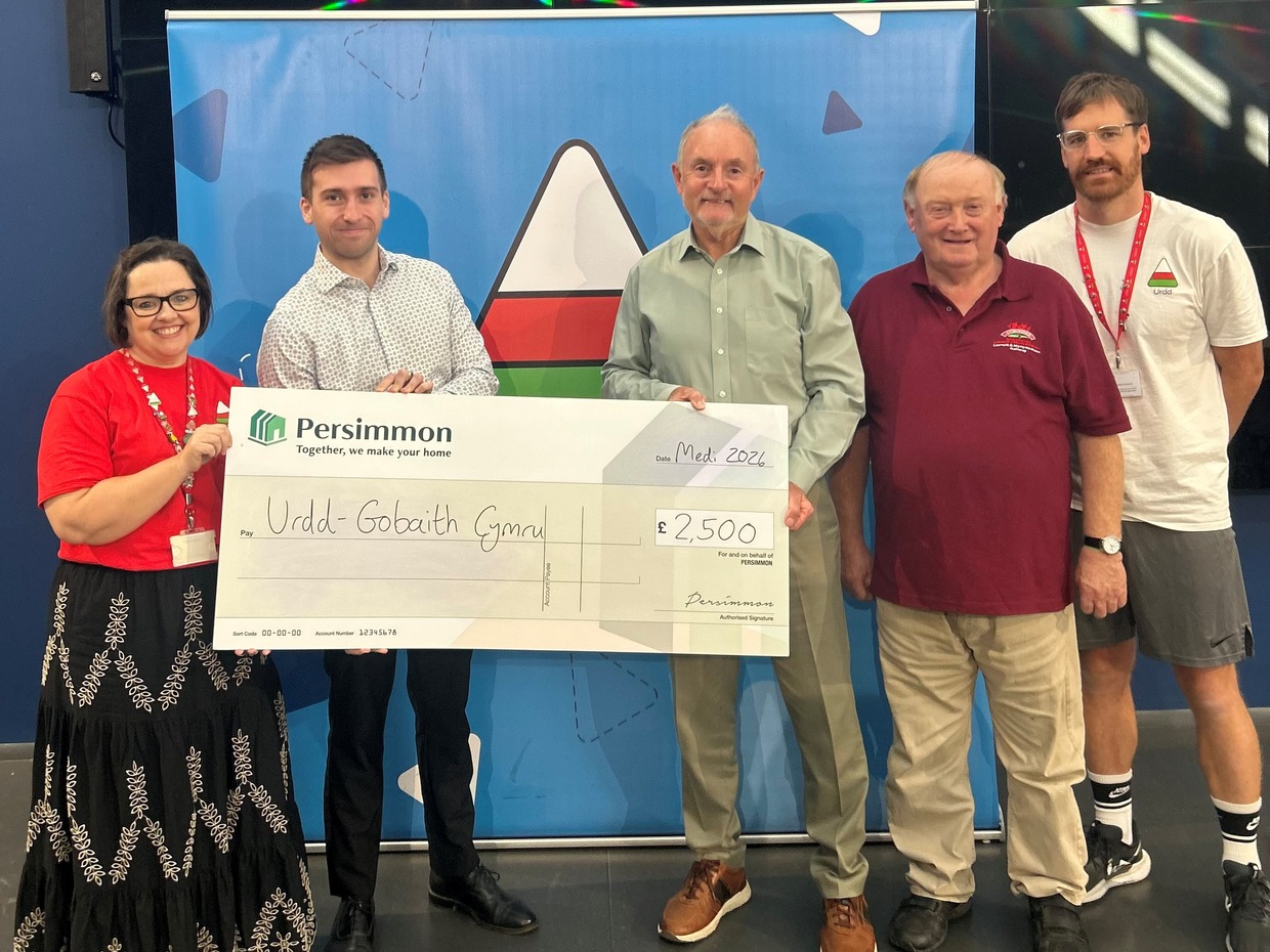 Five people standing in front of an Urdd banner holding a large Persimmon cheque made out to Urdd Gobaith Cymru for £2,500