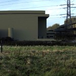 Photomontage showing large windowless data centre buildings and a multi-storey structure rising behind a hedgerow and rough grassland, with an electricity pylon between them