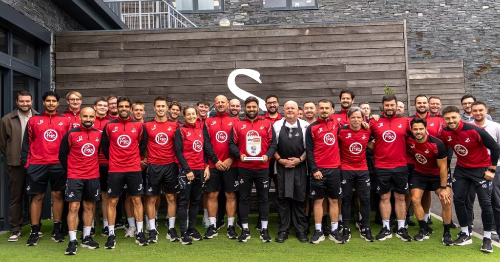 Vitor Matos holding the Sky Bet Championship Manager of the Month award, surrounded by Swansea City coaching and support staff at the training ground