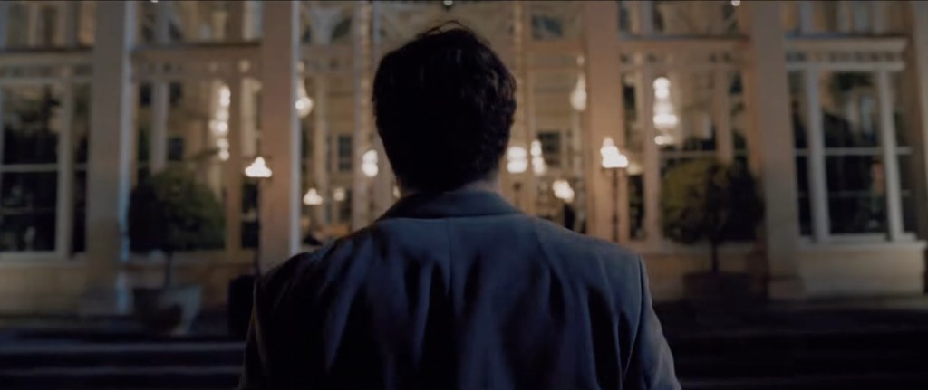 A man in a suit seen from behind, walking towards a grand country house lit up at night