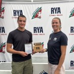 Wilf Tovey, a teenager in a dark T-shirt, holds a framed Impact Awards winner certificate and wooden RYA Cymru Wales trophy beside Sarah McGovern in front of an RYA Cymru Wales and Welsh Sailing backdrop