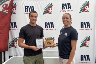 Wilf Tovey, a teenager in a dark T-shirt, holds a framed Impact Awards winner certificate and wooden RYA Cymru Wales trophy beside Sarah McGovern in front of an RYA Cymru Wales and Welsh Sailing backdrop