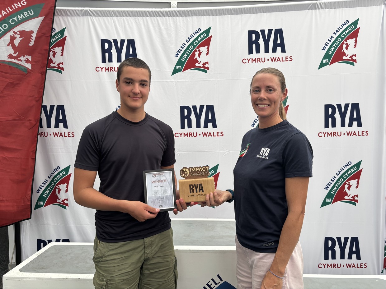 Wilf Tovey, a teenager in a dark T-shirt, holds a framed Impact Awards winner certificate and wooden RYA Cymru Wales trophy beside Sarah McGovern in front of an RYA Cymru Wales and Welsh Sailing backdrop