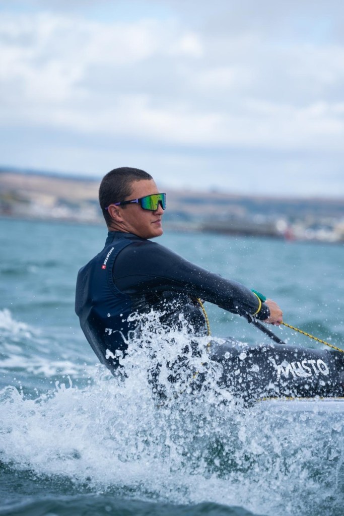 Close-up of Wilf Tovey in mirrored sunglasses and a wetsuit, mainsheet in hand, spray flying off the bow