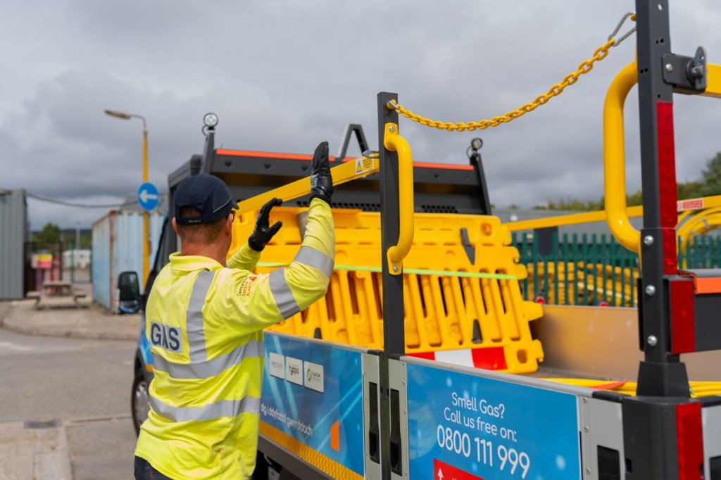 A Wales & West Utilities worker in a GAS hi-vis top loads yellow barriers onto the drop-side hydrogen van