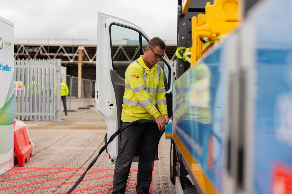 A worker connects the hydrogen refuelling hose to the white trial van beside its open cab door