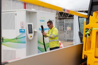 A worker in hi-vis holds the refuelling nozzle at a white hyQube hydrogen refueller, with the trial van behind