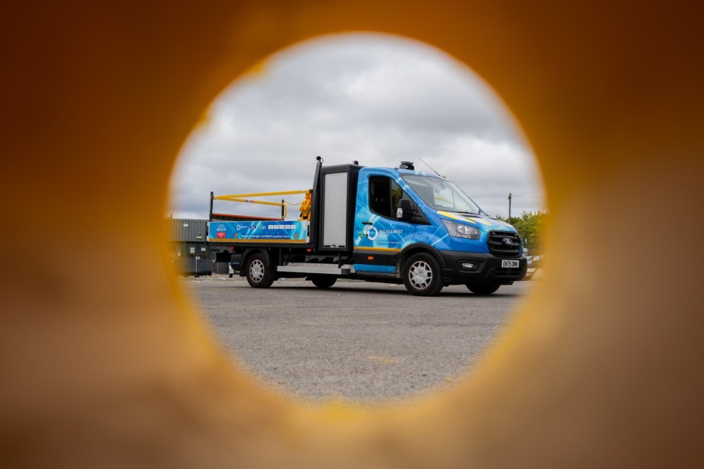 The blue-liveried Wales & West Utilities hydrogen Transit seen through the round hole of a yellow barrier