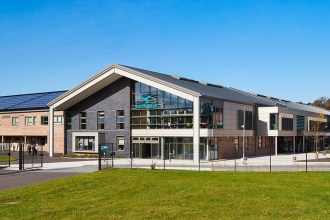 The modern brick and slate front of Ysgol Bro Dinefwr in Llandeilo under a blue sky, with solar panels on the roof and pupils crossing the grounds