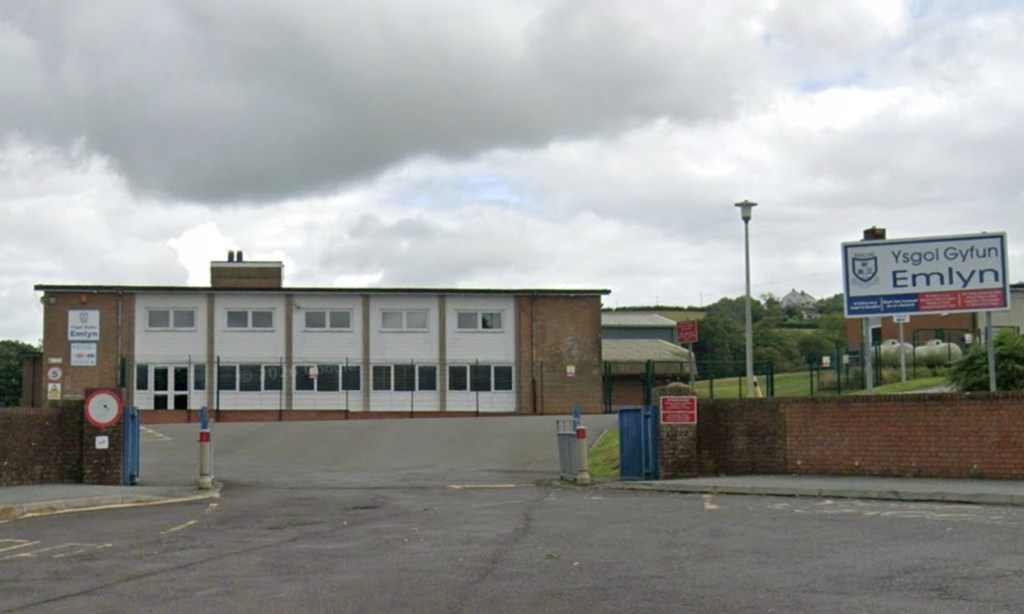 The entrance to Ysgol Gyfun Emlyn, a two-storey white and brick school building behind a gate, with the school's blue and white sign on the right
