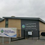 The front of Ysgol Gymraeg Ystalyfera, a brick and dark-panelled school building behind black fencing, with the school's welcome sign in front and the valley hillside behind