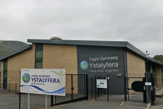 The front of Ysgol Gymraeg Ystalyfera, a brick and dark-panelled school building behind black fencing, with the school's welcome sign in front and the valley hillside behind