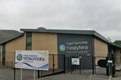 The front of Ysgol Gymraeg Ystalyfera, a brick and dark-panelled school building behind black fencing, with the school's welcome sign in front and the valley hillside behind