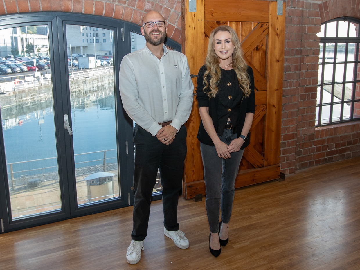 Alix Charles and James Smale-Davies standing in an empty brick-walled office unit at the J-Shed, with the dock visible through arched windows