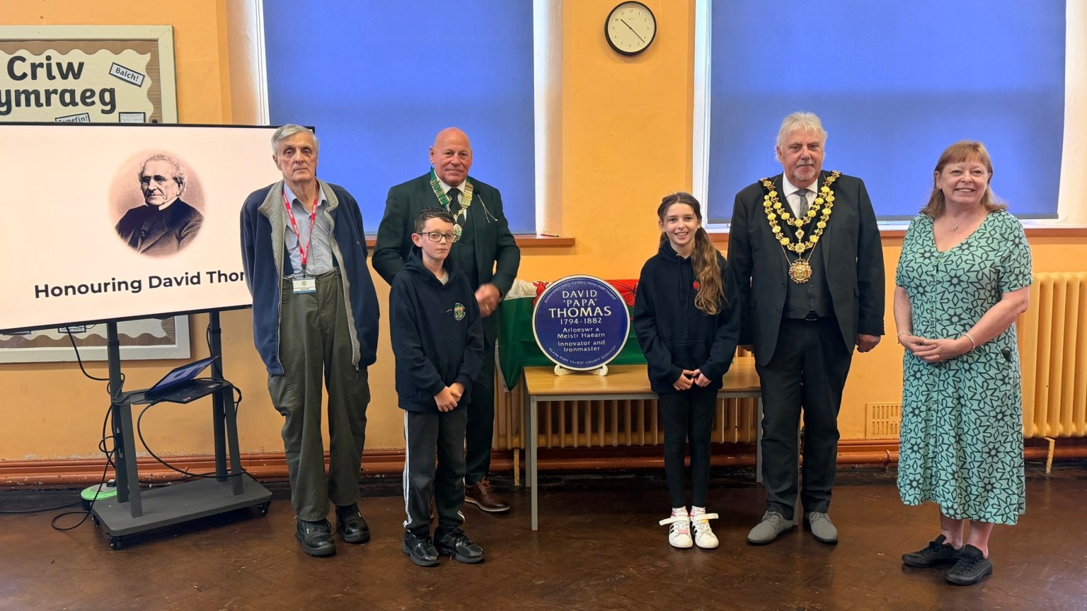 Two pupils and guests beside the blue plaque for David 'Papa' Thomas in a school hall, with his portrait on a screen