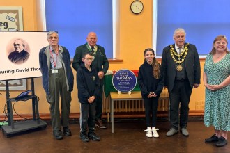 Two pupils and guests beside the blue plaque for David 'Papa' Thomas in a school hall, with his portrait on a screen