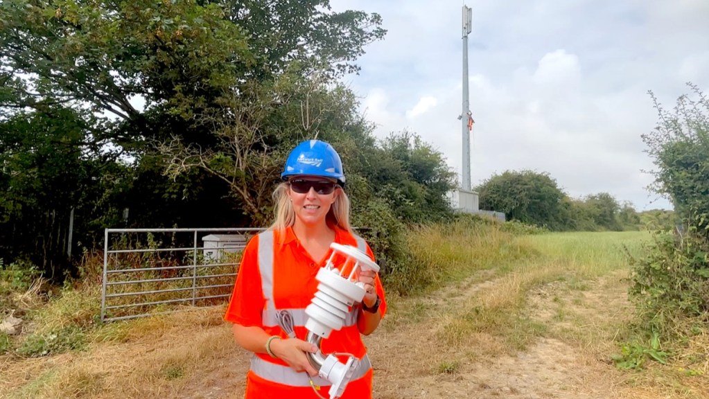 Amanda Williams of Network Rail, in orange hi-vis and a blue hard hat, holds a white weather station unit on a farm track with a railway radio mast behind her