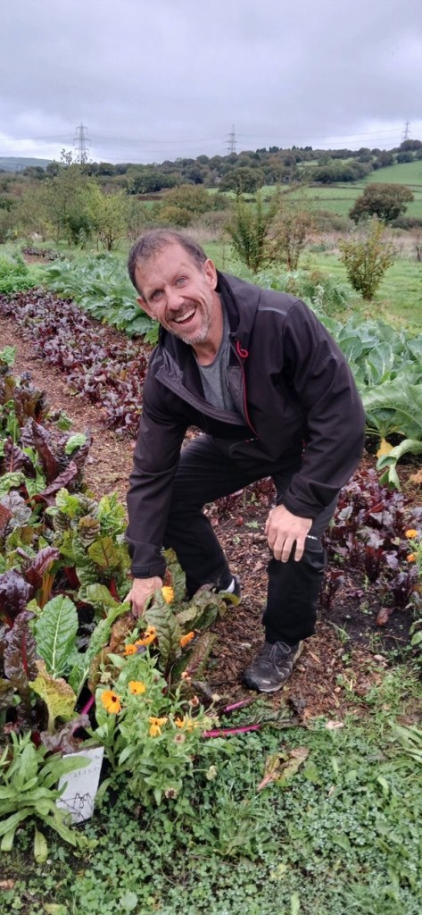 Andrew Jenkins crouching among rows of chard and marigolds at Cae Felin, smiling at the camera with open countryside and pylons behind