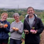 Traitors star Andrew Jenkins holding freshly harvested vegetables alongside two Cae Felin volunteers holding squashes and onions, with the farm and open countryside behind them