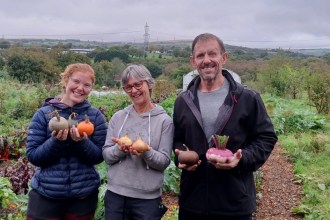 Traitors star Andrew Jenkins holding freshly harvested vegetables alongside two Cae Felin volunteers holding squashes and onions, with the farm and open countryside behind them