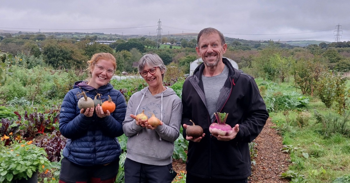 Traitors star Andrew Jenkins holding freshly harvested vegetables alongside two Cae Felin volunteers holding squashes and onions, with the farm and open countryside behind them