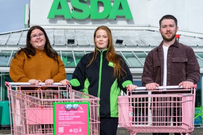 Three people with two pink shopping trolleys in front of an Asda store, one trolley carrying a pink poster asking 'Have you checked your chest?'