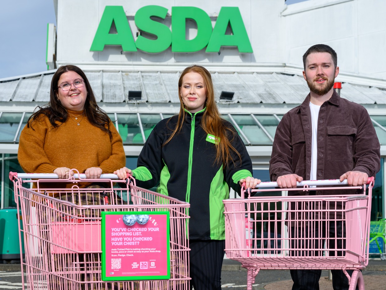 Three people with two pink shopping trolleys in front of an Asda store, one trolley carrying a pink poster asking 'Have you checked your chest?'