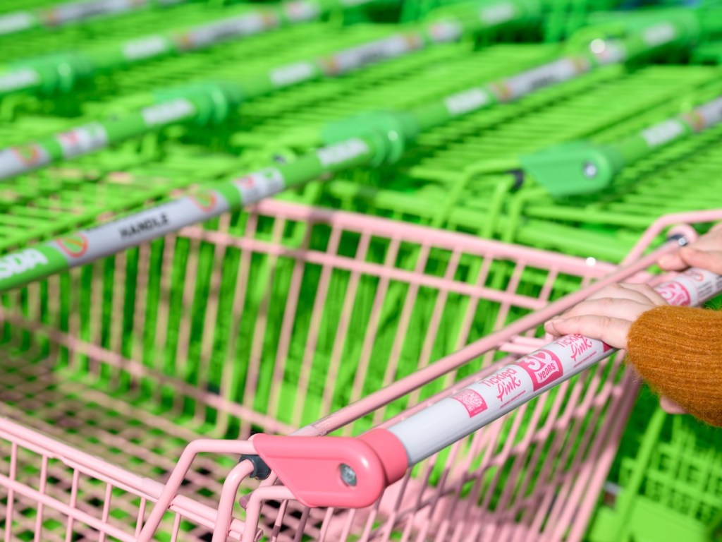 Close-up of hands on a pink Asda trolley handle printed with the Tickled Pink 30 years logo and a QR code, green trolleys behind