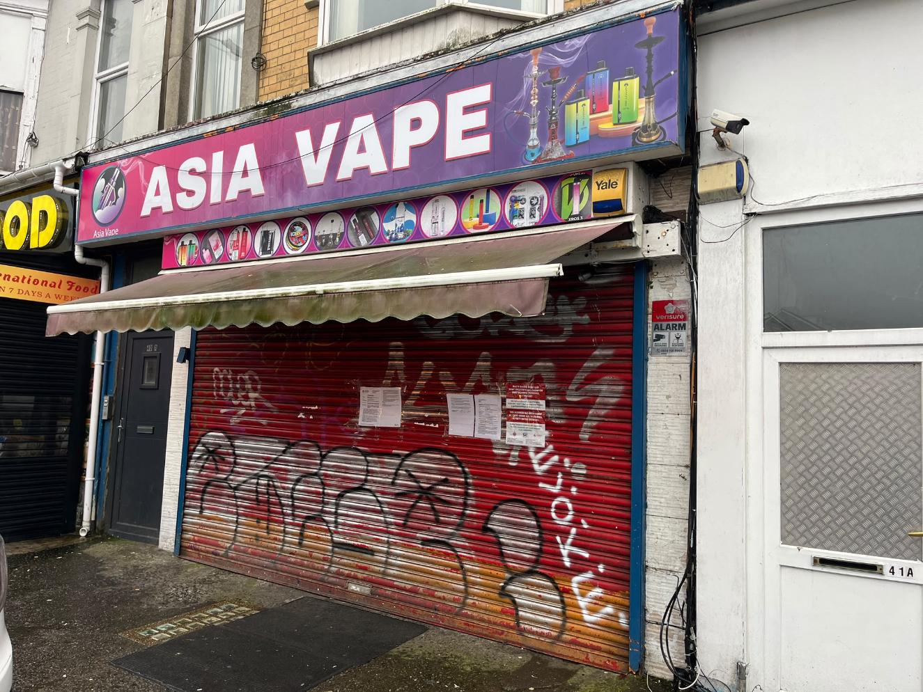 The red graffiti-covered shutter of Asia Vape on St Helen's Road, Swansea, with court closure notices taped to it under the shop's purple and pink sign