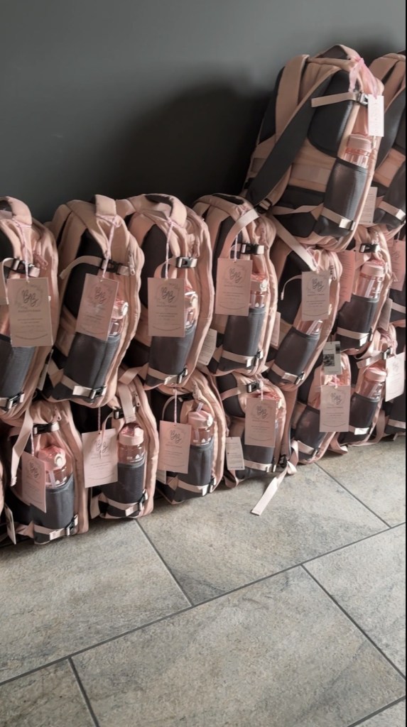 Rows of packed pink backpacks with water bottles and Bailey's Bags tags stacked against a wall