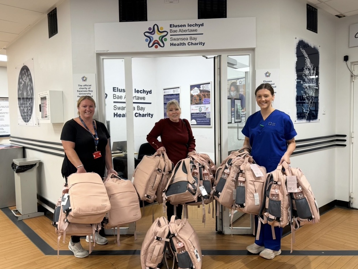 Maria Bailey and two others holding armfuls of pink Bailey's Bags backpacks outside the Swansea Bay Health Charity office