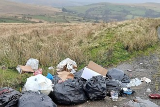 Black bin bags, cardboard boxes and plastic bottles dumped on the verge of a single-track moorland road, with green fields and hills behind