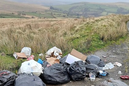 Black bin bags, cardboard boxes and plastic bottles dumped on the verge of a single-track moorland road, with green fields and hills behind