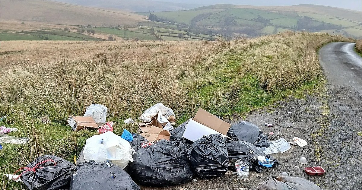 Black bin bags, cardboard boxes and plastic bottles dumped on the verge of a single-track moorland road, with green fields and hills behind