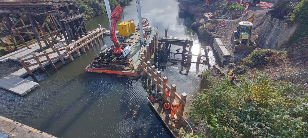 Workers in orange overalls in a small boat beside new timber fender rails in the River Tawe, with a crane barge and a digger on the bank