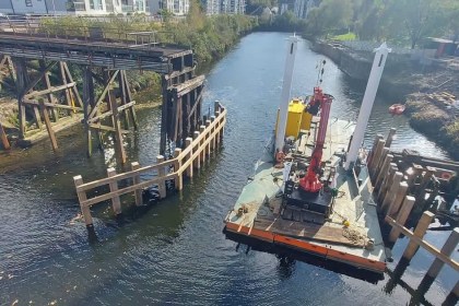 The old timber trestles of the Bascule Bridge standing in the River Tawe at Landore, with a crane barge and new timber fender rails alongside and riverside flats behind