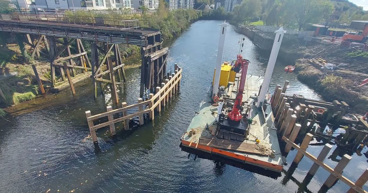 The old timber trestles of the Bascule Bridge standing in the River Tawe at Landore, with a crane barge and new timber fender rails alongside and riverside flats behind