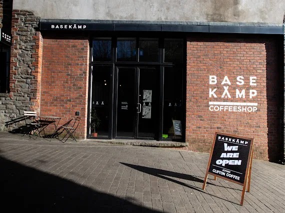 The red-brick frontage of Basekamp coffee shop on Kings Lane at Urban Village, Swansea, with its black doors and a We Are Open sign on the pavement