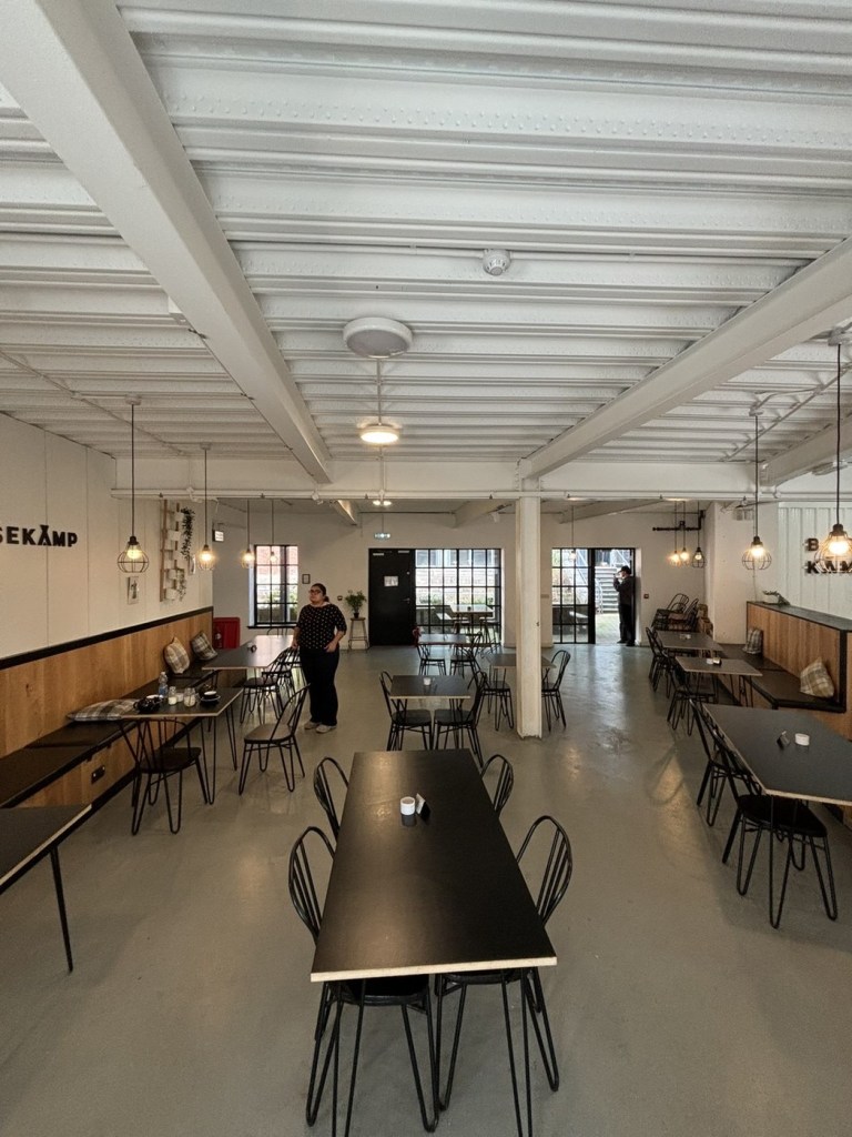 Inside Basekamp coffee shop on Kings Lane at Urban Village, Swansea: black tables and wire chairs under a white steel ceiling with pendant lights