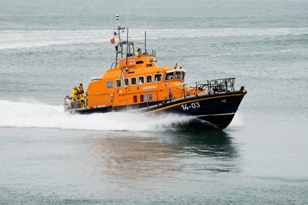 Fishguard RNLI's Trent class lifeboat Blue Peter VII, orange and navy with 14-03 on the bow, powering through grey sea with crew on the aft deck