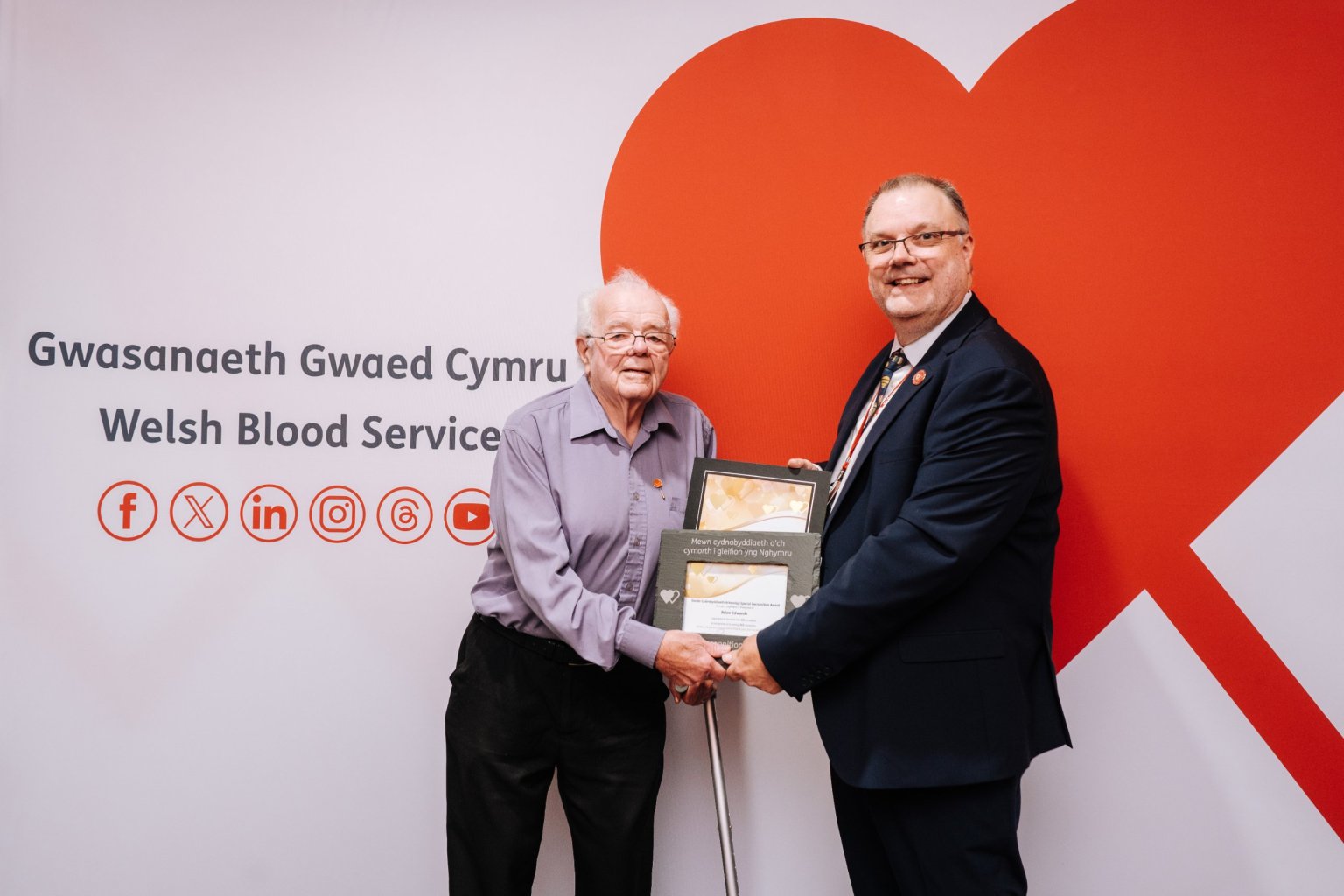 Brian Edwards, 83, holding a slate award with Peter Richardson of the Welsh Blood Service in front of a red heart backdrop