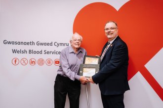 Brian Edwards, 83, holding a slate award with Peter Richardson of the Welsh Blood Service in front of a red heart backdrop