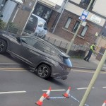 A grey SUV against a wall on Coity Road, Bridgend, with police tape and cones across the road and an officer by the Ty Ogwr building