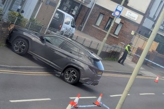 A grey SUV against a wall on Coity Road, Bridgend, with police tape and cones across the road and an officer by the Ty Ogwr building