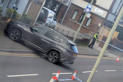 A grey SUV against a wall on Coity Road, Bridgend, with police tape and cones across the road and an officer by the Ty Ogwr building