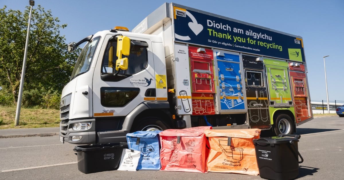 A Carmarthenshire Council recycling lorry with coloured compartments, parked behind blue, red and orange reusable sacks, a white battery sack and black food caddies