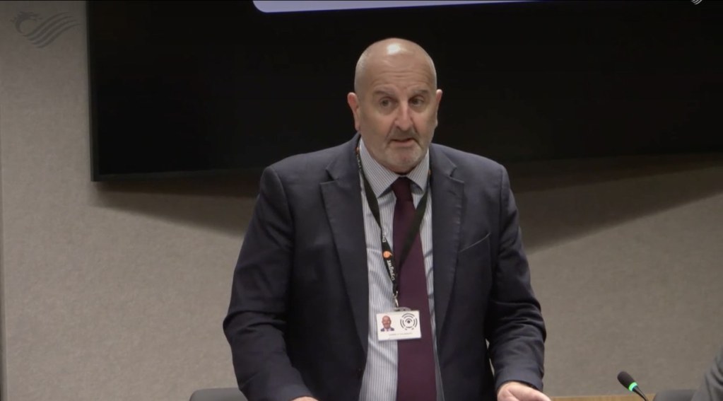 Carmelo Colasanto, in a navy suit with a Senedd lanyard, speaking in the Senedd chamber