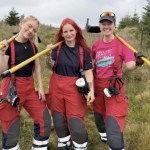 Three female firefighters in red fire trousers standing on moorland with yellow fire beaters over their shoulders, Cathryn Moore on the left