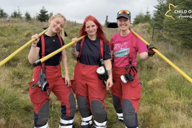 Three female firefighters in red fire trousers standing on moorland with yellow fire beaters over their shoulders, Cathryn Moore on the left