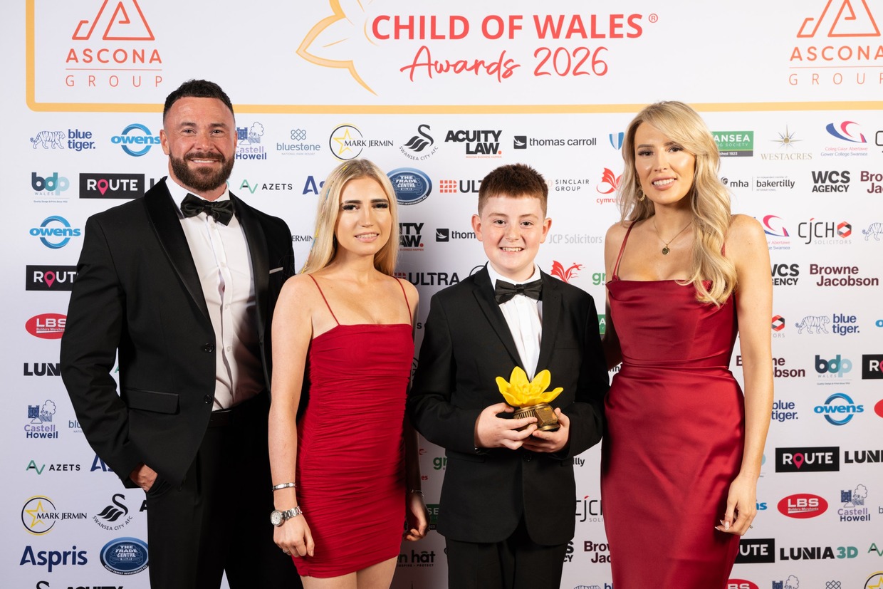 Cian Evans in a black suit and bow tie holding a yellow daffodil trophy, with his dad, Alex, and family in front of the Child of Wales Awards 2026 board